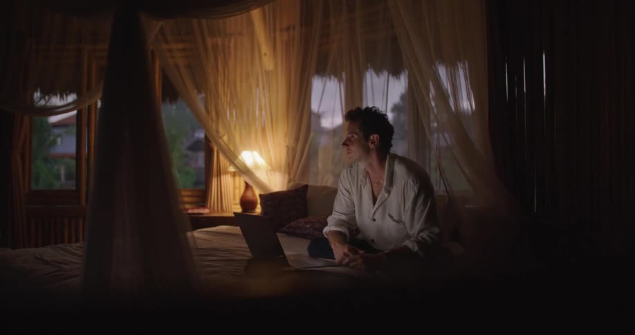 A man is sitting on his bed typing on a laptop computer and stops to look around as he is thinking and then types more