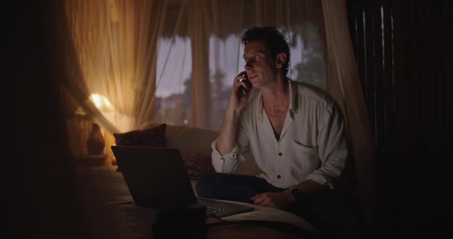 A man is sitting on his bed laughing as he talks on the phone and types on a laptop computer