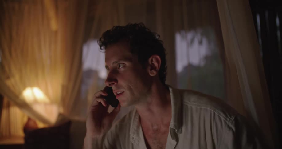 Man with white shirt sits on bed talking on smartphone in dim lit bedroom