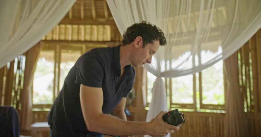 A man examines a camera before packing it into a carrying case while standing in a bedroom during the day