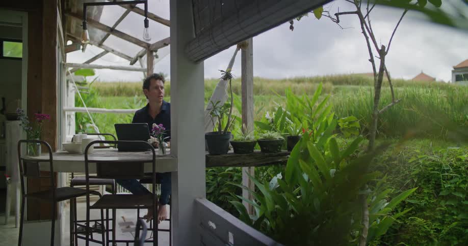 A man types on a laptop while sitting at a table on the patio of a house in a large field during the day