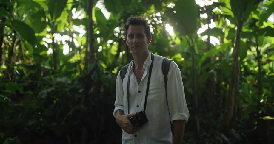 A man is standing outside surrounded by tropical plants and holding the camera that is hanging around his neck and he smiles