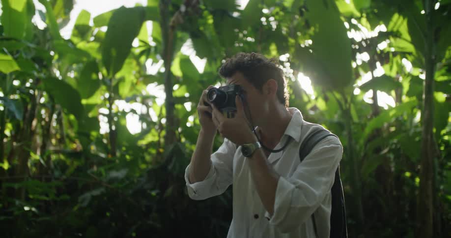 Man with white shirt backpack and wristwatch takes photographs with camera in jungle