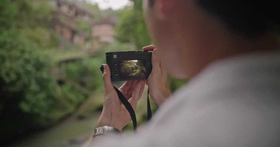 A man is standing outside holding his camera and looking at the scene in the viewfinder on the back of the camera