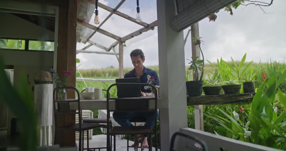 A man is sitting at a table outside on a patio typing on a laptop computer