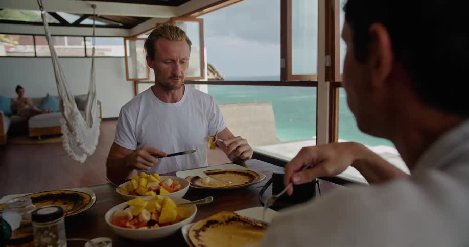 Woman sits on couch as two male friends eat and talk at tablenear window with view of water