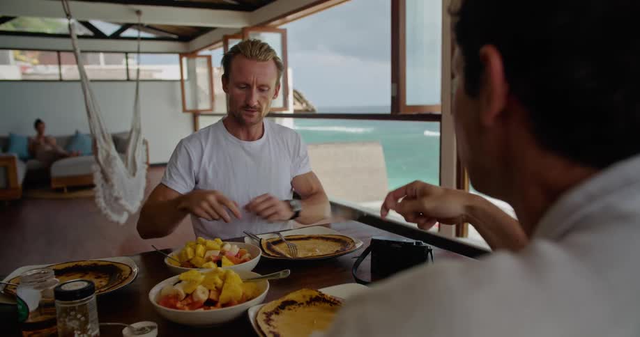 Two men sitting at table near window with view of ocean talk and eat while a woman sits on a couch in the background