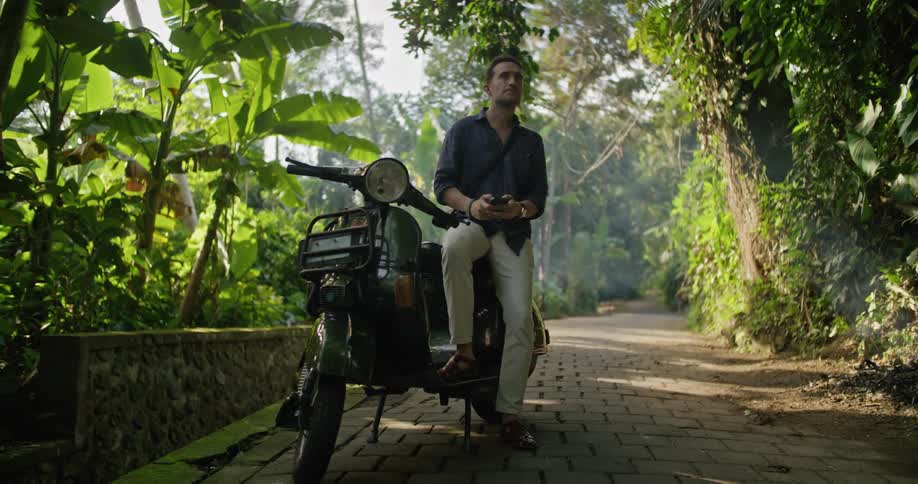 A man is sitting on his motorbike on a cobblestone road using his cell phone and looking around