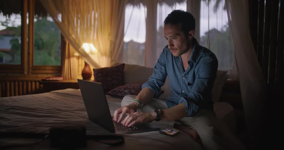 Man with blue shirtbrown pants and wristwatch sitting on bed types on laptop in dim lit bedroom