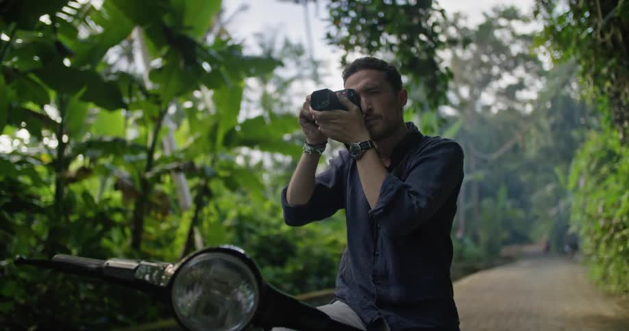 A man is sitting on his motorbike on a cobblestone walkway surrounded by tropical plants as he lifts his camera and takes a picture