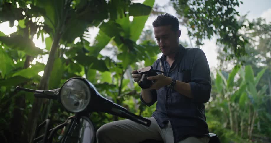 A man is sitting in on his motorcycle in a rainforest and he lifts his camera to take a picture