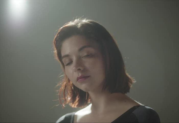 A woman is sitting under a light as cameras flash and she moves her hair with her hand