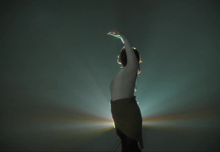A woman is dancing and moving her arms twisting her body as a light shines brightly behind her
