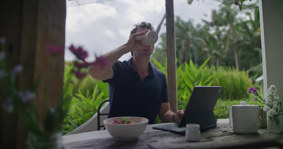 Young man works on a laptop while eating breakfast on the porch of a tropical house