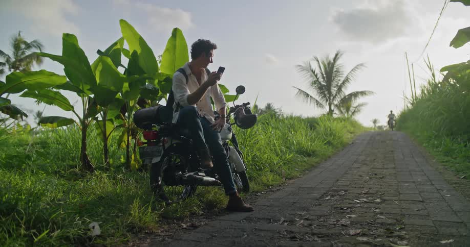 A man is sitting on his motorbike on the side of a cobblestone road using his cell phone as another man on a motorbike rides by