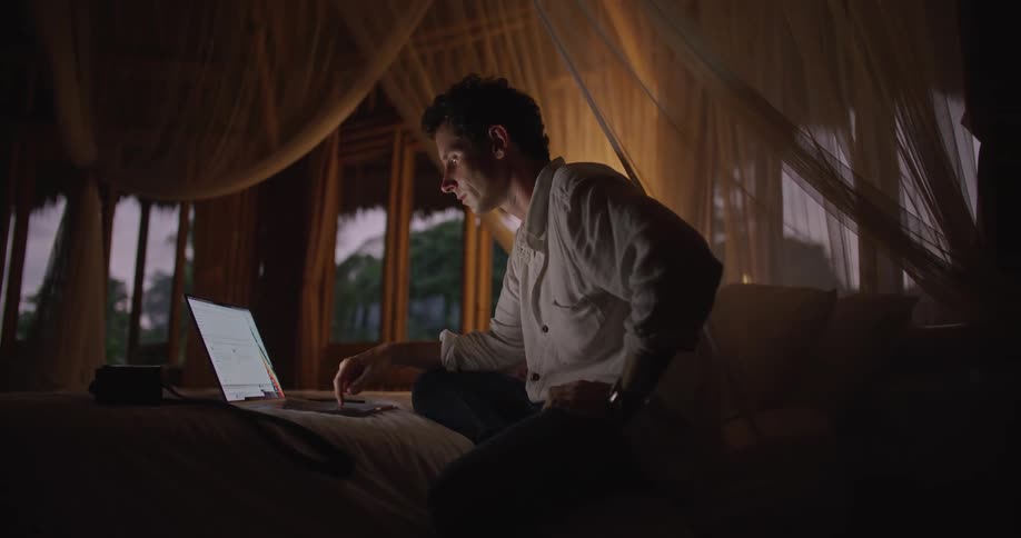 Man with blue shirt and brown pants sits on bed working and closing laptop in dim lit room
