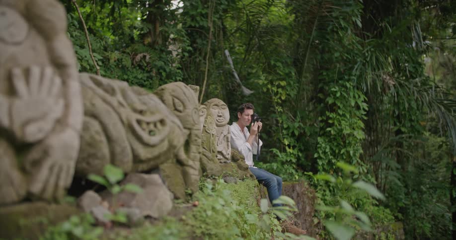 A man is sitting on a wall next to tiki statues as he lifts his camera to his face and takes a picture