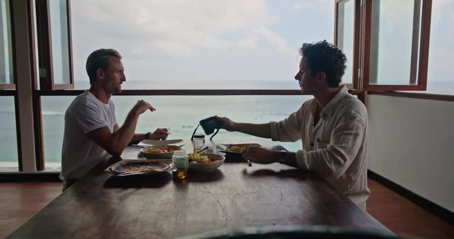 A man checks his camera while eating together with another man in a dining room overlooking the ocean
