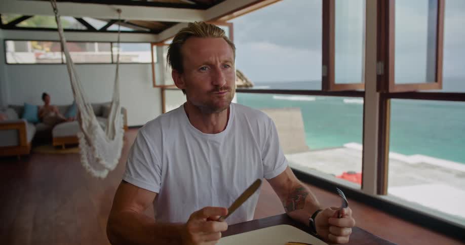 Man smiles while eating with another at table near window showing the beach
