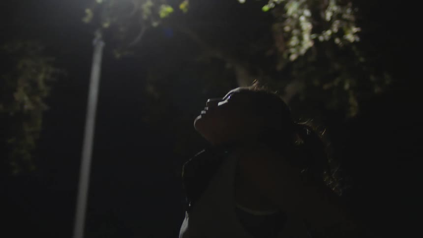 A woman stretches her back while standing outdoors under a street light at night