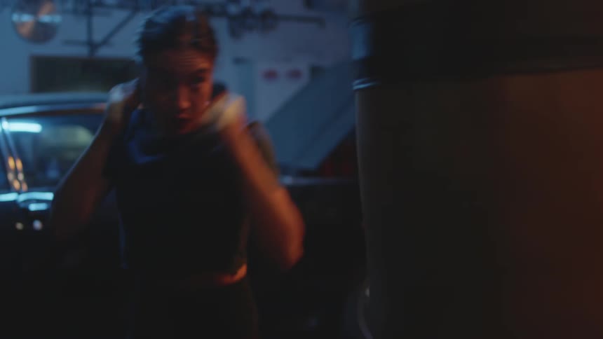 A woman strikes a punching bag in a dimly lit garage
