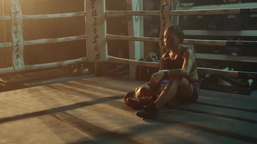 Young female boxer sits tiredly on the floor of a boxing ring at evening