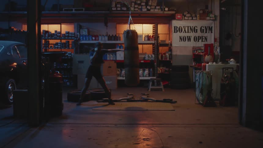 Woman works with punching bag hung near parked car and full shelves of garage