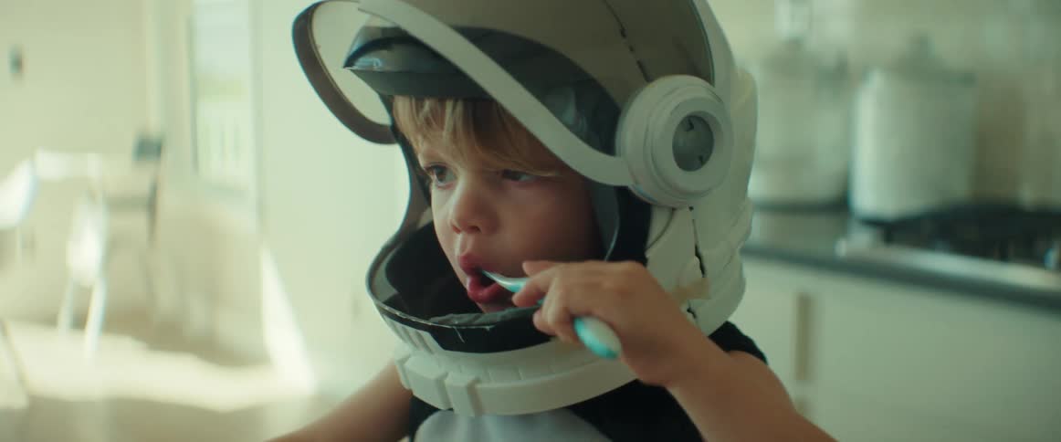 A young child wearing a toy astronaut helmet brushes his teeth