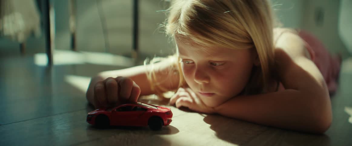 The sun shines upon a little girl lying on the floor playing with a toy car