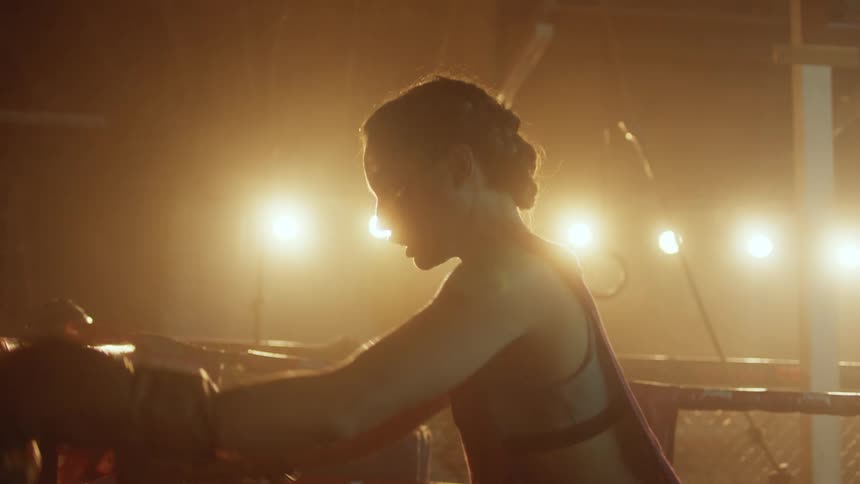 Female boxer standing in ring stretches her back while another woman sits nearby on outside of ring
