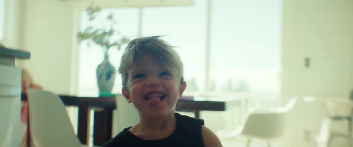 A pair of young boys chase the camera through the living room and kitchen of a house during the day
