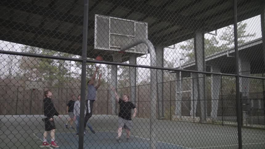 A group of young men play basketball on an outdoors basketball court during the day