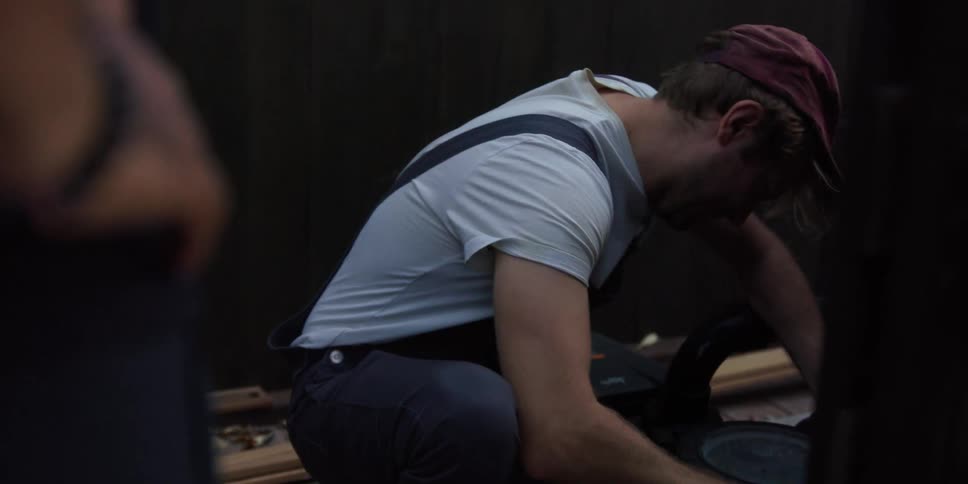 A man wearing suspenders and a cap on his head is squatting and reaching down with both arms