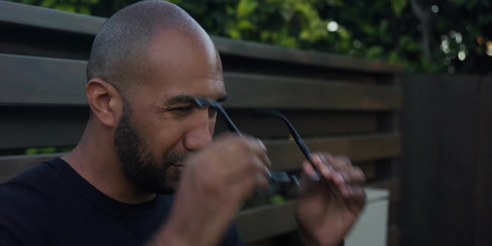 A man is standing outside near a fence and he takes off his glasses and looks at them then puts them back on his face