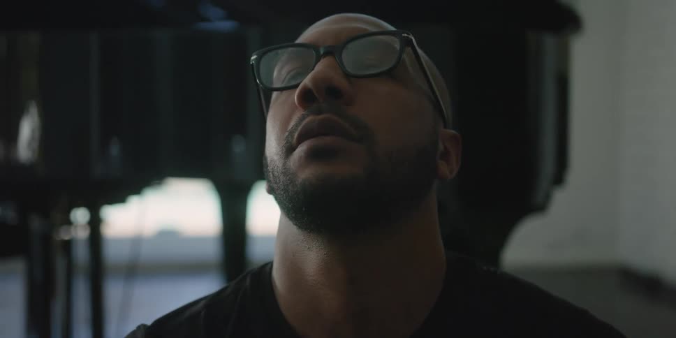 Bearded man wearing glasses with a towel over her neck sitting on the floor near a grand piano