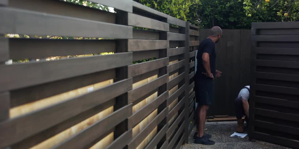 Man standing near a fence in the backyard talking to a worker crouching behind a wall