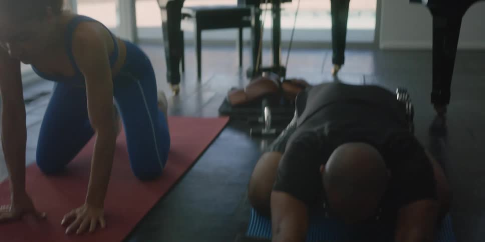 Man with shaved head beard glasses and black t shirt stretches his body next to woman with ponytail blue sports bra and pants in front of grand piano