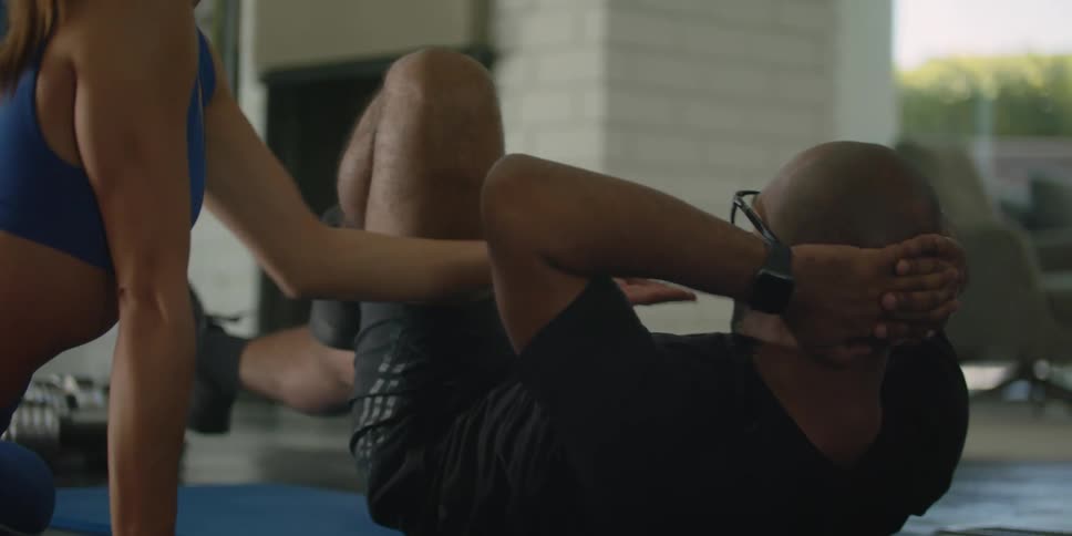 A man does bicycle crunches on an exercise mat while being instructed by a personal trainer