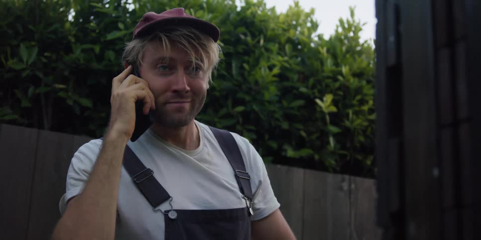 A man in overalls answers his smartphone while standing outdoors during the day