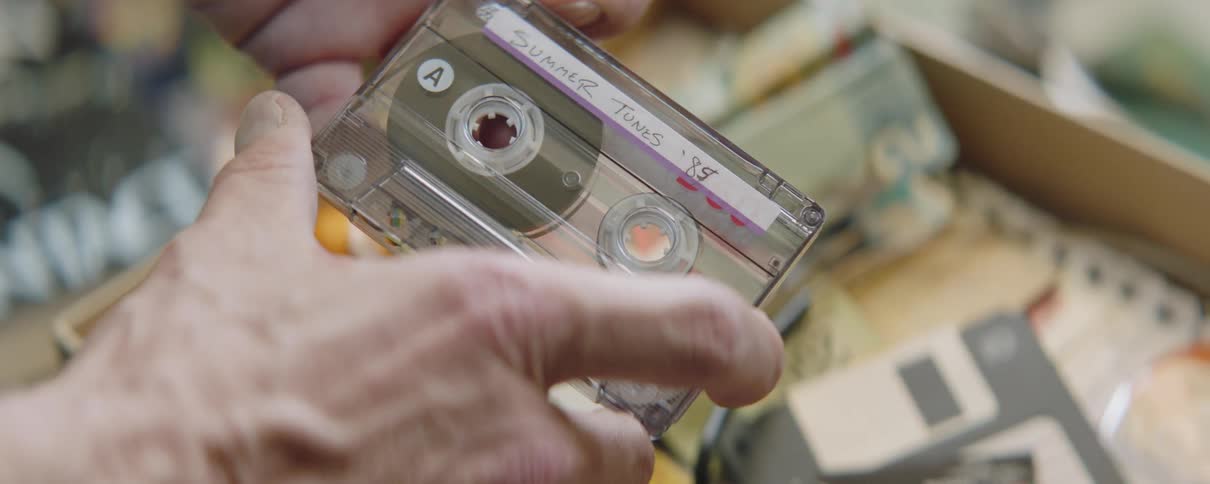 Older male examines labeled cassette tape near box of items that include a floppy disk
