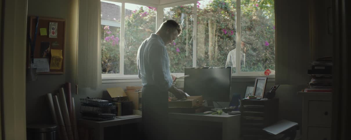 A man is standing over a desk with a box in front of computer monitor and he is looking though letters in the box as the sunlight shines through the windows