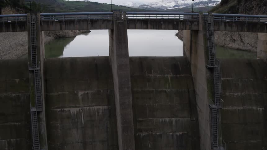 Panning up the walls of a large dam to see the reservoir and mountains behind it