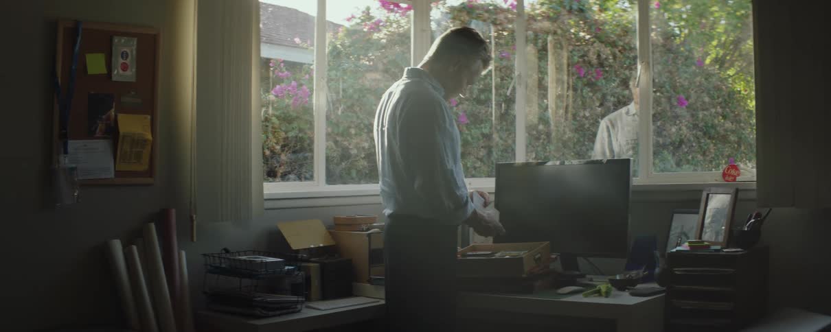 A man is standing over a desk with a box in front of computer monitor and he is looking though things in the box as the sunlight shines through the windows
