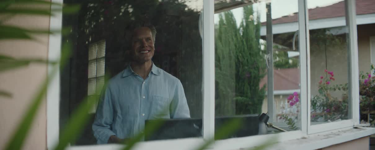 A man stands in his home in front of a computer and smiles as he looks out the window