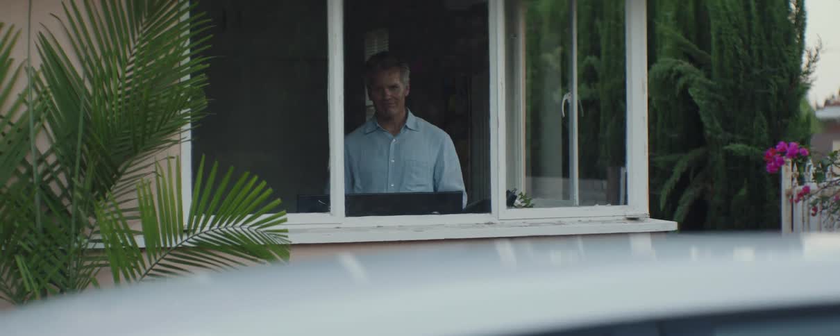 A man is inside the house looking out the window as a teenage girl gets into a car in the driveway