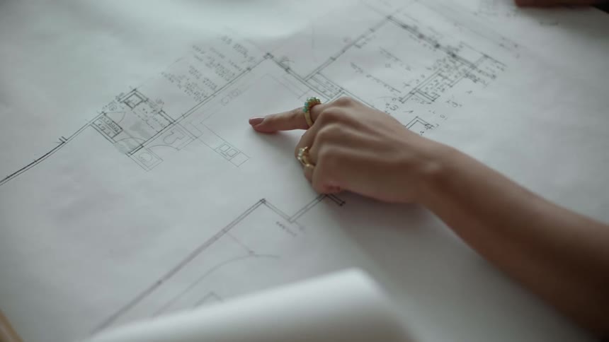 Drafting plans are on a table as a woman moves her hand pointing to an area a man places his hand on top of hers and holds her hand