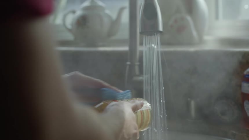 Young man doing dishes in the kitchen sink uses steaming hot water to clean a glass