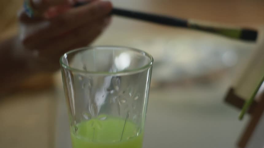 A painter using watercolors dips the brush on a glass with water before painting on canvas