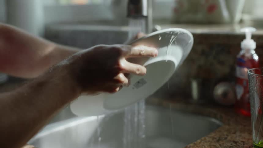 Young person doing dishes in the kitchen sink uses steaming hot water to clean a plate