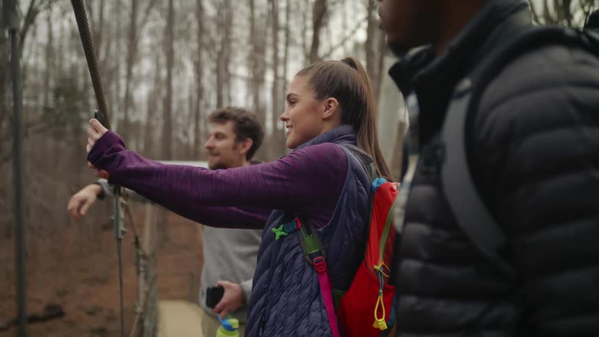 Woman with ponytail red backpack purple sweater and dark blue sweater takes selfie between two men on forest bridge